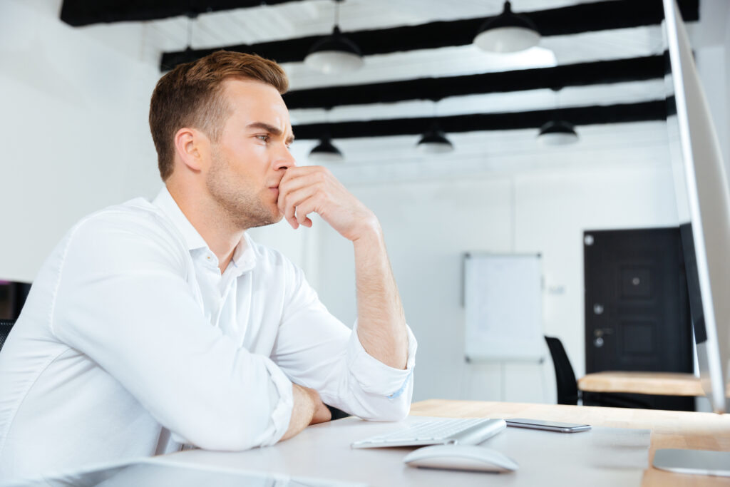 Pensive young businesman working with computer and thinking at workplace